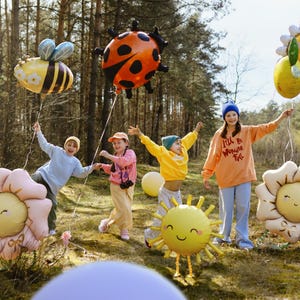 May include: Four children in a forest setting holding colorful balloons shaped like a ladybug, bee, sun, and flowers. The balloons have happy faces and the children are smiling. One child is wearing a blue hat and a peach colored sweatshirt with the text "I'll be Wonder-ful" on it.