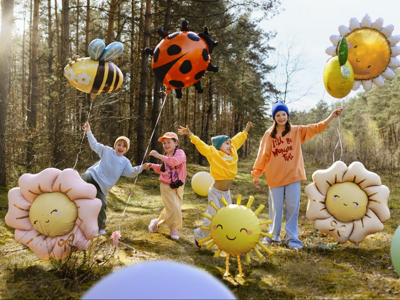 May include: Four children in a forest setting holding colorful balloons shaped like a ladybug, bee, sun, and flowers. The balloons have happy faces and the children are smiling. One child is wearing a blue hat and a peach colored sweatshirt with the text "I'll be Wonder-ful" on it.