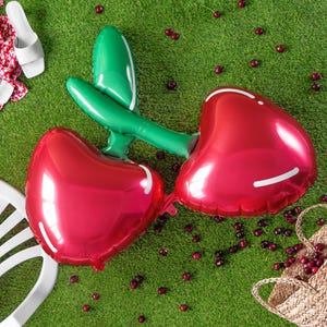 A single foil balloon shaped like two connected red cherries with green stems, lying on green artificial grass. Scattered real cherries surround the balloon, along with summer items like a white plastic chair, white sandals, a red floral fabric, and a woven straw bag.