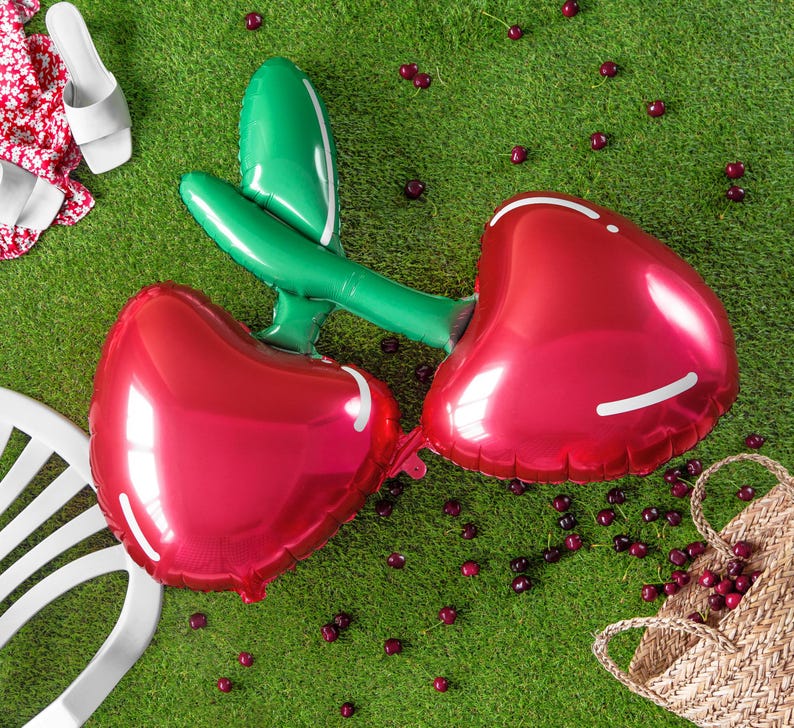 A single foil balloon shaped like two connected red cherries with green stems, lying on green artificial grass. Scattered real cherries surround the balloon, along with summer items like a white plastic chair, white sandals, a red floral fabric, and a woven straw bag.