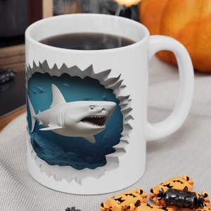 Puede incluir: Taza de café de cerámica blanca con una imagen 3D de un gran tiburón blanco rompiendo la superficie de la taza. El tiburón nada en agua azul sobre un fondo blanco.