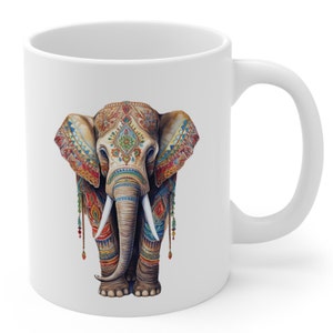 Puede incluir: Taza de cerámica blanca con una ilustración colorida de un elefante decorado con intrincados patrones y joyas. El elefante está de frente con la trompa baja.
