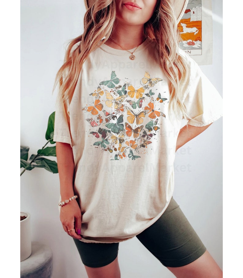 May include: A beige oversized t-shirt with a graphic print of colorful butterflies. The shirt is worn with dark green biker shorts.