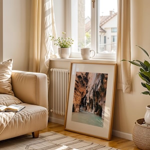 May include: A framed print of a canyon landscape with a turquoise river running through it. The print is hanging on a wall in a living room with a beige couch, a potted plant, and a window with white curtains.