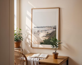 May include: A framed photograph of a beach scene with people on the sand and a pier in the distance. The photo is hanging on a wall in a room with a wooden desk and a potted plant.