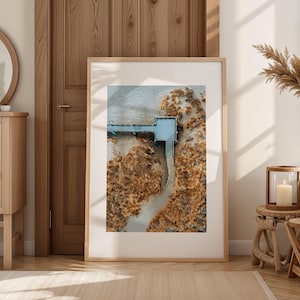 Könnte beinhalten: Ein gerahmter Druck mit einer Luftaufnahme eines Piers, der sich in ein Gewässer erstreckt und von goldbrauner Vegetation umgeben ist. Der Pier hat eine hellblaue Struktur. Das Kunstwerk wird in einem Raum mit Holzmöbeln und -dekoration ausgestellt.