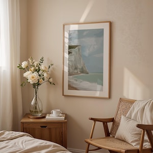 May include: A white wooden bedside table with a drawer and a white ceramic mug on top. A clear glass vase with white flowers sits on the bedside table. A framed print of a seascape with a white cliff hangs on the wall behind the bedside table. A wooden chair with a white throw blanket sits in the corner of the room.