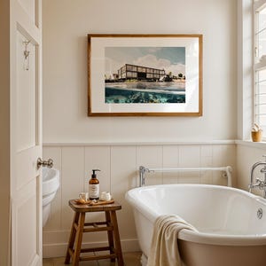 May include: A bathroom scene featuring a framed picture of a building over water, a white clawfoot tub, and a small wooden stool with toiletries. The room has a neutral color palette with a window and a partially open door.