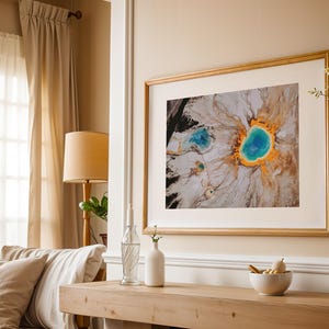 May include: A framed photograph of a colorful aerial view of a geothermal hot spring with a blue and green center surrounded by orange and white. The photograph is hanging on a beige wall above a wooden table.