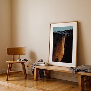 May include: A framed photograph of a waterfall cascading down a cliff face. The photograph is hanging on a light beige wall above a wooden bench. A wooden chair is in the foreground.