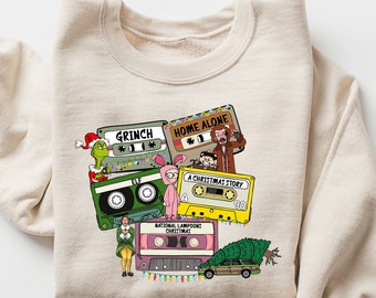 May include: A beige sweatshirt with a graphic print of cassette tapes featuring characters from popular Christmas movies, including "The Grinch", "Home Alone", "A Christmas Story", and "National Lampoon's Christmas Vacation".