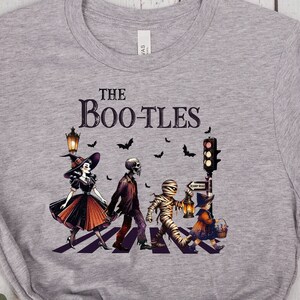 Op de afbeelding: Een heather grijze t-shirt met een Halloween-thema grafisch ontwerp. Het ontwerp toont verklede figuren, waaronder een heks, een skelet en een mummie, die een zebrapad oversteken. De tekst "The Boo-tles" staat boven de figuren.