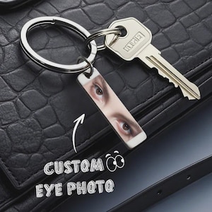 May include: A silver keychain with a rectangular tag featuring a custom eye photo. The keychain is next to a silver key. The words "CUSTOM EYE PHOTO" are written in white with an arrow pointing to the keychain.