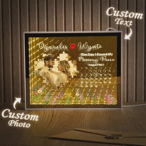 Op de afbeelding: Een gepersonaliseerde verlichte fotolijst met een foto van een stel en de tekst "The Day I Found My Missing Piece". De lijst bevat een kalender voor augustus 2023 en bloemmotieven. De lijst is rechthoekig en heeft een warm, gloeiend licht.