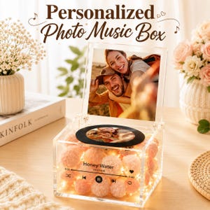 May include: A clear acrylic music box featuring a hinged lid with a photo of a couple. The front panel displays the text Honey Water by VICETONE with a music player interface. The interior is filled with pink pom-poms and warm fairy lights.