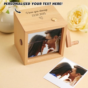 Könnte beinhalten: Eine Holz-Spieluhr mit einem Foto eines Paares, einer Kurbel und dem Text "I love you always 22.07.2025" mit einem Herzsymbol. Auf der Oberseite der Box steht der Text "PERSONALISIERE DEINEN TEXT HIER!". Ein goldfarbener Knopf ist sichtbar.