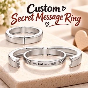 Puede incluir: Dos anillos de metal plateado con un acabado liso y pulido. Un anillo está cerrado, mientras que el otro está parcialmente abierto, revelando un grabado interior que dice: "You had me at hello". El texto "Custom Secret Message Ring" aparece arriba.