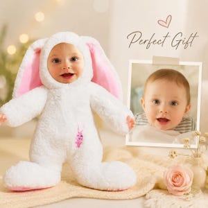 May include: A white plush baby jumpsuit with long rabbit ears and a pink interior lining. The suit features a small embroidered pink rabbit on the chest. Next to the suit is a framed photograph of a baby and the text Perfect Gift with a heart icon.