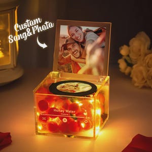 Pode incluir: Uma caixa de música de acrílico transparente com uma foto e um pequeno disco. A caixa é iluminada com luzes brancas quentes e preenchida com esferas vermelhas. O texto "Custom Song & Photo" é visível. A caixa tem uma foto de um casal.