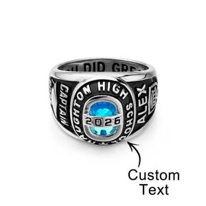 May include: A silver-colored class ring with a black band. The ring features a large, oval, light blue stone at the center, with the year "2026" visible. The ring is engraved with the words "HOUGHTON HIGH SCHOOL" and "CAPTAIN".