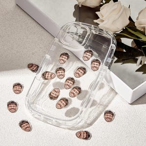 Könnte beinhalten: Eine transparente Handyhülle mit einer Reihe kleiner Aufkleber, die ein lächelndes Gesicht mit Brille zeigen. Die Aufkleber sind um die Hülle und auf der Oberfläche verteilt. Die Hülle liegt auf einer weißen Oberfläche mit weißen Blumen.