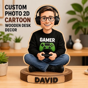 Op de afbeelding: Een op maat gemaakt 2D houten bureau-decoratie met een cartoonillustratie van een persoon met een zwarte koptelefoon, bril en een zwart sweatshirt met de tekst GAMER en een groene controller. De houten voet toont de naam DAVID in het zwart.