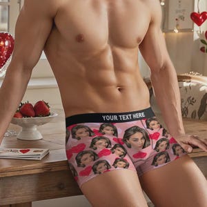 May include: Pink boxer shorts featuring a pattern of faces and red hearts. The black waistband displays the text "YOUR TEXT HERE" in white. The shorts appear to be made from a soft, stretchy fabric.