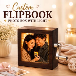 Op de afbeelding: Een vierkante flipbook fotobox van donker hout met een ingebouwd licht en een zilveren slinger aan de zijkant. De voorkant toont een foto van een glimlachend stel. De tekst erboven luidt Custom FLIPBOOK PHOTO BOX WITH LIGHT.