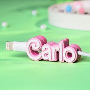 May include: Pink cable organizer with the name "Carlo" in white letters. The organizer is made of soft, flexible material and is designed to keep cables tidy and organized.