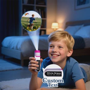 May include: A boy smiles while holding a white and pink projector flashlight. The flashlight projects an image of a boy flying a kite with a dog. The flashlight has the text "Olivia Grace March 14, 2025" and "Custom Text".