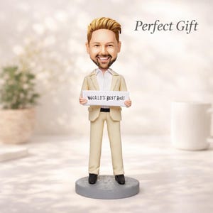 May include: A bobblehead figurine of a smiling man in a tan suit holding a sign that reads "WORLD'S BEST BOSS." The figurine has a light skin tone, blond hair, and is standing on a gray circular base. The words "Perfect Gift" are written above the figurine.
