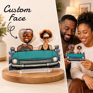 May include: A custom acrylic display featuring a cartoon illustration of two people in a teal convertible car. The design includes cutouts for faces, with the text Custom Face and an arrow pointing to the head area. The piece stands on a clear base.
