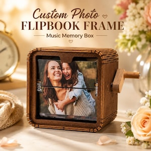 Puede incluir: Un marco de madera marrón oscuro para libro animado con una manivela en el lateral. La parte frontal muestra una fotografía de una mujer y un niño. El texto superior dice Custom Photo FLIPBOOK FRAME Music Memory Box en una fuente serif marrón.