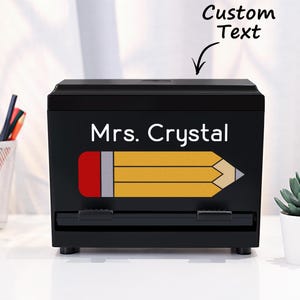 Può includere: Un organizer da scrivania nero con la scritta "Mrs. Crystal" e una grafica a matita. Il design della matita presenta una gomma rossa, una ghiera argentata e un corpo giallo. Le parole "Custom Text" sono sopra l'organizer.