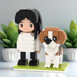 Può includere: Una figura in mattoni di una persona con i capelli neri e un cappotto bianco, in piedi accanto a un cane in mattoni. Il cane è marrone, bianco e nero. Entrambe le figure sono su una base verde, con piante verdi sullo sfondo.