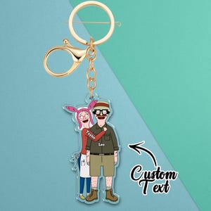 May include: A gold-coloured keyring featuring a cartoon image of two people. The image shows a woman with pink bunny ears hugging a man. The woman's name "Taylor" is on her shirt, and the man's name "Leo" is on his shirt. The words "Custom Text" are also on the keyring.