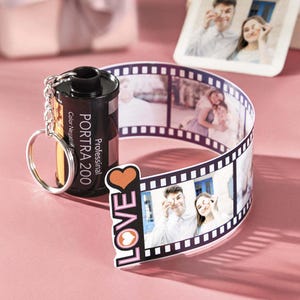 May include: A black film canister keychain with a white label that reads "Professional Portra 200 Color Negative" and a pink "LOVE" charm with hearts. The keychain is attached to a strip of film with photos of a couple.