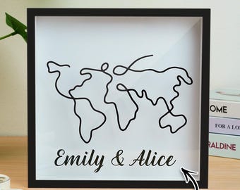 May include: A framed wall art piece featuring a minimalist black line drawing of a world map. The names "Emily & Alice" are printed in a script font below the map. The frame is black, and the background is white. The words "Custom Text" are in a speech bubble.