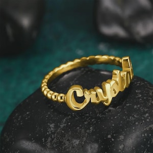 May include: A gold-colored ring with the name "Crystal" in a cursive font. The ring band has a beaded design. The ring is displayed on a dark, textured surface.