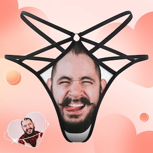 May include: Black thong underwear with a printed image of a man's face, complete with a mustache and tongue out. The underwear has a distinctive strappy design with black straps.