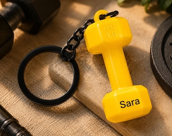 May include: A bright yellow dumbbell-shaped keychain featuring a black metal ring and chain. The dumbbell has the text 1KG LB embossed on the top and the name Sara printed in black on the bottom weight section, creating a distinct fitness accessory.