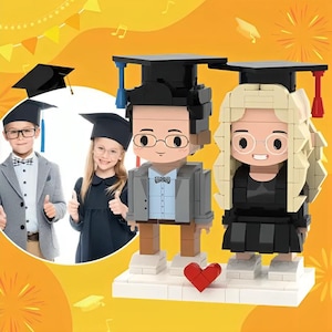 May include: Two Lego minifigures wearing graduation caps and gowns. The figures are standing on a white base with a red heart in the center. The figures are a boy with brown hair and a girl with blonde hair.