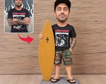 May include: A custom bobblehead figurine of a person wearing a black t-shirt with the text LEARN TO FORGET, camouflage shorts, and a black cap. The figurine holds a gold surfboard and stands on a circular gray base, modeled after the person in the photo.