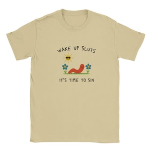 以下が含まれることがあります： 「WAKE UP SLUTS, IT'S TIME TO SIN」のテキストが入ったタン色のTシャツ。サングラスをかけた笑顔の太陽、赤いミミズ、青い花が描かれています。このシャツは柔らかい素材で作られています。