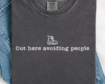 May include: A dark gray t-shirt with the text "Out here avoiding people" and a small white graphic of a hiking boot. The shirt is folded on top of a pair of blue jeans. The shirt is labeled "Pepper".