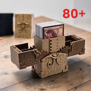 Puede incluir: Una caja para barajas de plástico color bronce con detalles de engranajes mecánicos y patrones jeroglíficos grabados, que contiene un mazo de cartas. La caja tiene un diseño abierto con compartimentos laterales y el texto rojo 80+.