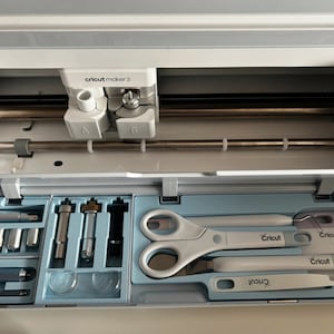Puede incluir: Una máquina Cricut Maker 3 abierta con la tapa abierta, revelando el compartimento de almacenamiento de herramientas. El compartimento contiene una variedad de herramientas, incluyendo un par de tijeras, un raspador, una herramienta de deshierbe y varias cuchillas. Las herramientas están etiquetadas con el nombre de la marca Cricut.