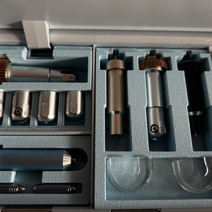 Op de afbeelding: Een opbergdoos met diverse metalen snij- en embosstools voor een knutselmachine. De set bevat zilveren en zwarte cilindrische behuizingen, sommige met messingkleurige tandwielen, en kleine puntige uiteinden met nummers zoals 02, 11, 31 en 41.