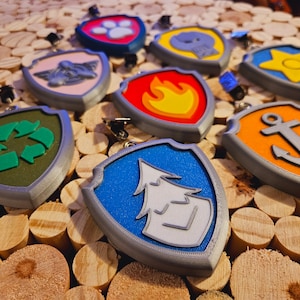 Könnte beinhalten: Ein Set aus zehn bunten Plastikabzeichen mit verschiedenen Designs, darunter ein Pfotenabdruck, ein Feuer, ein Recyclingsymbol, ein Baum, ein Stern, ein Anker und ein Schädel. Die Abzeichen sind alle silberfarben und haben einen Clip auf der Rückseite.