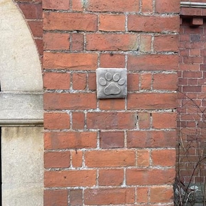 May include: A square, grey stone plaque featuring a paw print is set into a red brick wall. The paw print is a detailed relief, showing the pads and toes. The brickwork is a mix of red and brown tones, with visible mortar.